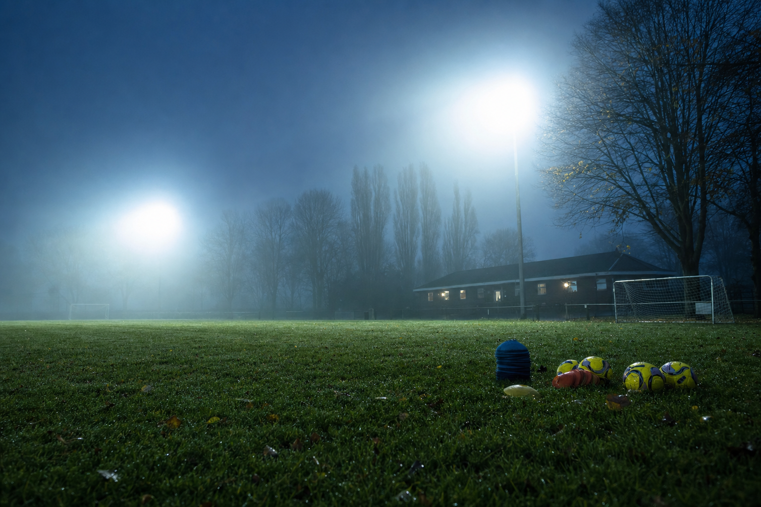 Amateurfußballplatz im Herbstnebel unter Flutlicht, Hütchen und Bälle auf dem nassen Rasen, im Hintergrund kahle Bäume und ein kleines Vereinsheim